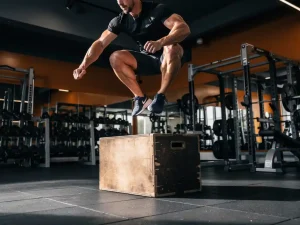 Atleet op maximale hoogte tijdens een explosieve box jump in moderne sportschool, dramatisch laag camerastandpunt benadrukt kracht.