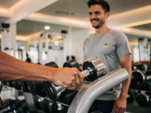 Beginner die een lichte dumbbell vasthoudt bij een gewichtenrek, met een trainer op de achtergrond in een moderne, goed verlichte sportschool.