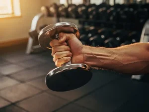 Gespierde onderarm met dumbbell tijdens bicepscurl, zweet op de huid, sportschool met gewichtsrekken op de achtergrond in warm goudkleurig licht.
