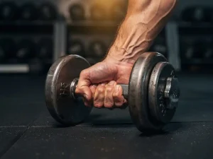 Gespierde onderarm met zichtbare aders die een zware ijzeren dumbbell curl uitvoert in een sportschool, met gewichtsrekken op de achtergrond.