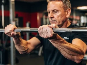 Middelbare persoon grijpt een halter tijdens krachttraining in moderne sportschool, met zichtbare aders en gefocuste blik.
