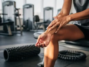 Persoon masseert pijnlijke onderarmspier op gymbank, met foam roller en weerstandsband ernaast, in moderne sportschool.