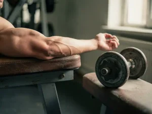 Verslapte biceps op een gymbank met een stoffige dumbbell op de achtergrond, symboliseert spierverlies door inactiviteit.