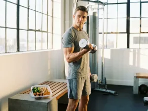 Gespierde atleet voert dumbbell curl uit in moderne gym, met maaltijdprep bakje met kip en groenten op nabijgelegen bank.