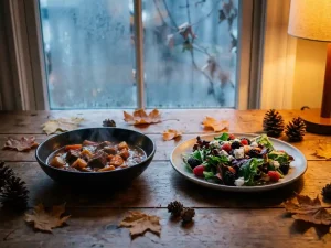 Houten eettafel met winterse stoofpot en zomerse salade, omringd door herfstbladeren, warm gouden licht, bevroren raam op achtergrond.